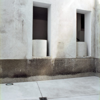 View from under the canopy, in the foreground the new concrete pavement, in the plastered wall two existing, transformed openings with cylindric concrete parapet elements - complicity of materials